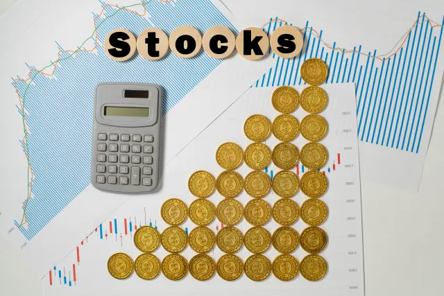 グラフの資料の上に階段状に並べた金色のコインと「Stocks」の木製文字、電卓